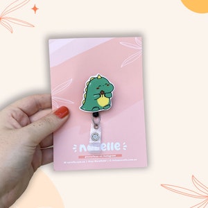 May include: A green dinosaur cartoon character with a pink heart on its chest, holding a brown chocolate bar. The dinosaur is attached to a white retractable badge holder with a silver clip. The background is a pink and white striped pattern with a white floral design. The text "norelle" is printed in black on the badge holder. The text "@norelleau on Instagram" is printed in black below the badge holder. The text "www.norelle.com.au | Etsy: NorelleAu | info@norelle.com.au" is printed in black below the Instagram text.