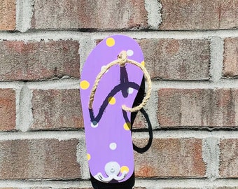 Puede incluir: Chancla morada con lunares amarillos y blancos, un pulpo blanco y un asa de cuerda. La chancla está montada en una pared de ladrillo.