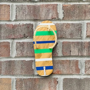 May include: A wooden flip-flop shaped wall hook with a rope loop. The flip-flop is painted with white, blue, and green stripes.