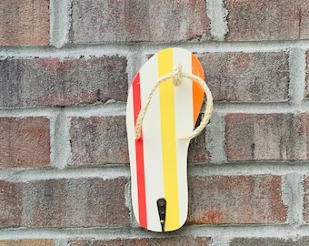 Puede incluir: Un gancho de pared decorativo con forma de chancla. La chancla tiene rayas verticales en rojo, amarillo y naranja. Un lazo de cuerda sirve de correa. El gancho está montado en una pared de ladrillo.