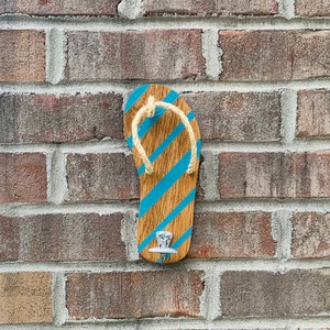 May include: Wooden flip-flop shaped wall hook with blue diagonal stripes and a rope strap. The hook is mounted on a brick wall, offering a beachy or coastal decor theme.