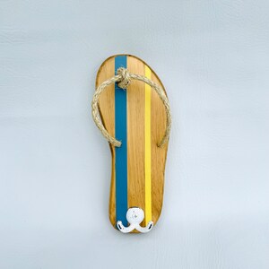 May include: Wooden flip-flop shaped wall hook with blue and yellow stripes and a rope detail. The hook has a white metal double hook.
