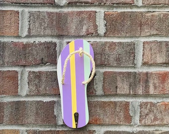 Puede incluir: Una chancla de color morado, amarillo y verde con una tira de cuerda colgada en una pared de ladrillo.