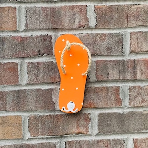 May include: Orange flip flop with white polka dots and a rope strap. The flip flop is hanging on a brick wall.
