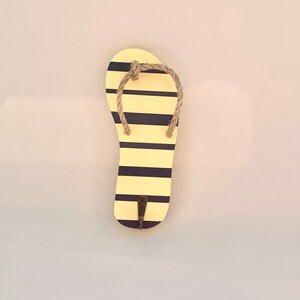 May include: A yellow and black striped flip flop shaped wall hook. The hook is made of wood and has a rope detail around the top.