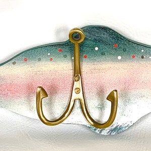 May include: A wooden fish-shaped wall hook with a gold-toned double hook. The fish is painted in shades of blue, green, pink, and white with black spots. The fish has a yellow eye.