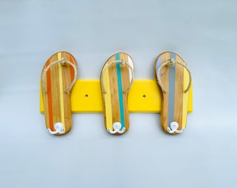 Puede incluir: Tres ganchos de madera con forma de chanclas montados en una tabla de madera pintada de amarillo. Las chanclas están pintadas con rayas en tonos naranja, amarillo, azul y gris.