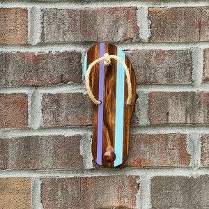 May include: A wooden flip-flop shaped hook painted in light blue and purple with a brown wood grain finish. The hook has a natural twine rope wrapped around it.