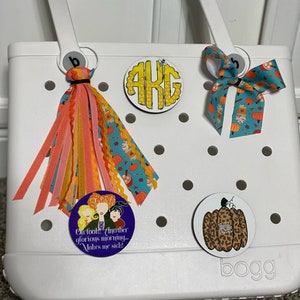 May include: White tote bag with a variety of round, colorful, fabric patches. The patches feature a monogram, a pumpkin, and a group of witches. The bag has a white handle and the word "BOGG" is printed on the bottom.