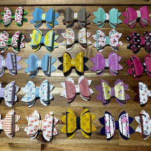 May include: A collection of 24 handmade hair bows in various colors and patterns. The bows are made of faux leather and feature a variety of designs, including floral, geometric, and abstract patterns.
