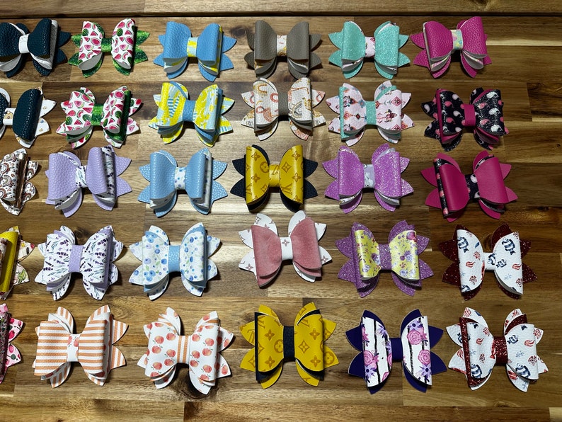 May include: A collection of 24 handmade hair bows in various colors and patterns. The bows are made of faux leather and feature a variety of designs, including floral, geometric, and abstract patterns.