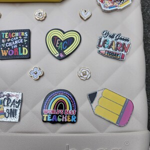 K&ouml;nnte beinhalten: Eine Sammlung dekorativer Charms und Anstecknadeln, darunter ein Bleistift, ein Herz und ein Regenbogen, mit Texten wie "TEACHERS CAN CHANGE THE WORLD" und "WORLD'S BEST TEACHER". Die Artikel sind in verschiedenen Farben und Designs gehalten, passend f&uuml;r Lehrer.