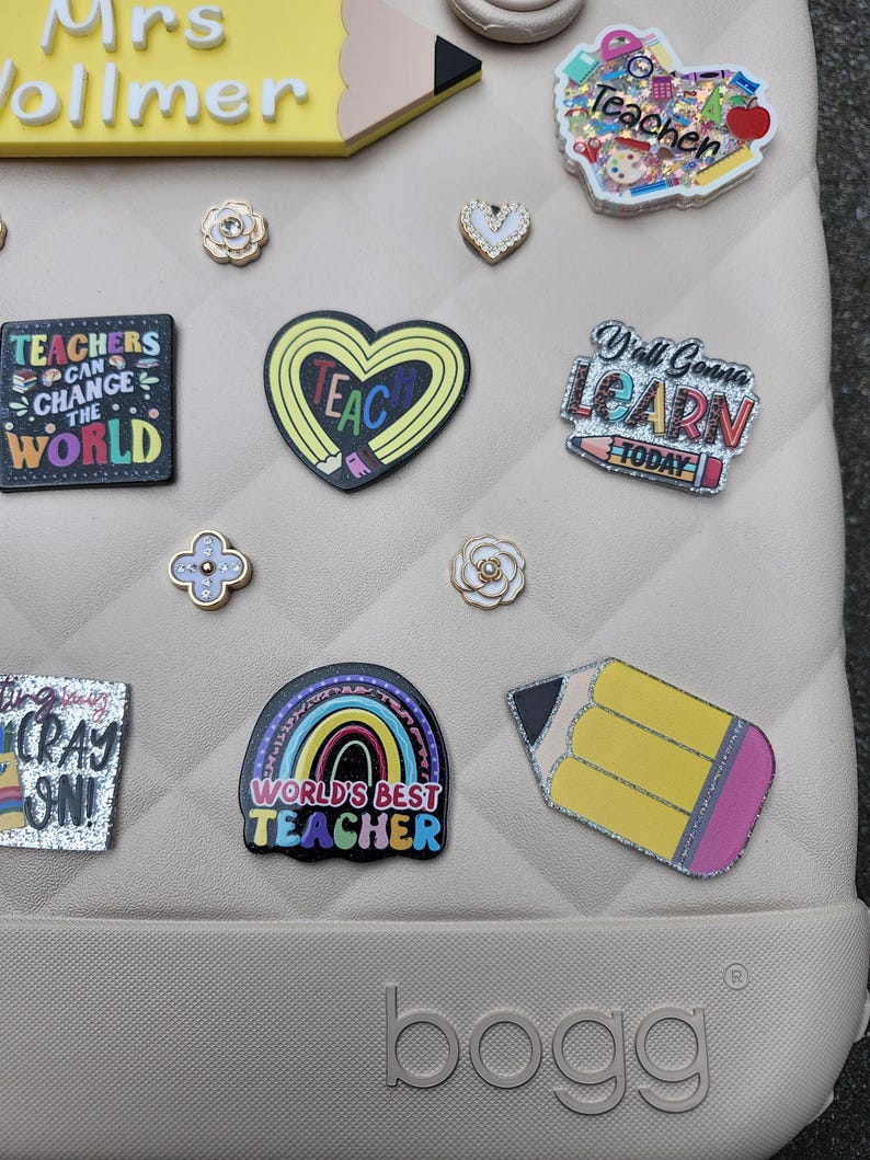 K&ouml;nnte beinhalten: Eine Sammlung dekorativer Charms und Anstecknadeln, darunter ein Bleistift, ein Herz und ein Regenbogen, mit Texten wie "TEACHERS CAN CHANGE THE WORLD" und "WORLD'S BEST TEACHER". Die Artikel sind in verschiedenen Farben und Designs gehalten, passend f&uuml;r Lehrer.