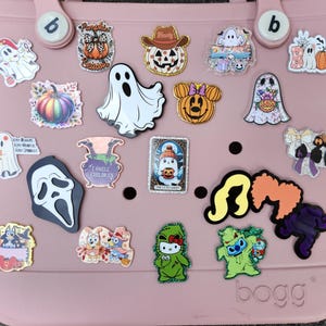 May include: A pink tote bag adorned with various decorative charms. The charms feature Halloween-themed designs like ghosts, pumpkins, and a Scream mask, alongside cartoon characters. The bag has two handles and the word "bogg" printed on the bottom.
