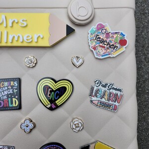 K&ouml;nnte beinhalten: Eine beige Tragetasche, verziert mit verschiedenen dekorativen Ansteckern. Zu den Ansteckern geh&ouml;ren ein gelber Bleistift, ein herzf&ouml;rmiger Anstecker mit dem Wort "TEACH" und Anstecker mit dem Text "Teacher", "Y'all Gonna LEARN TODAY" und "WORLD'S BEST TEACHER".