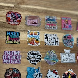 May include: A collection of colorful stickers with various sayings and designs. The stickers feature glitter, skulls, and cartoon characters. Some of the sayings include "Just Peachy", "Have the Day You Deserve", "I'm Fine It's Just My Face", "On My Husband's Last Nerve", "I Kinda Don't Care", "Normal People Scare Me", "The Day You Deserve", "Stay Tuned For My Next Dramatic Episode", "Have the Day You Deserve", "Don't Be a Salty Bitch".
