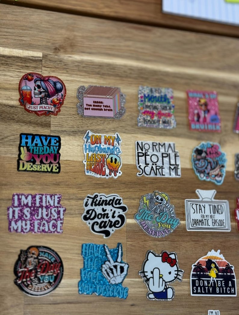 May include: A collection of colorful stickers with various sayings and designs. The stickers feature glitter, skulls, and cartoon characters. Some of the sayings include "Just Peachy", "Have the Day You Deserve", "I'm Fine It's Just My Face", "On My Husband's Last Nerve", "I Kinda Don't Care", "Normal People Scare Me", "The Day You Deserve", "Stay Tuned For My Next Dramatic Episode", "Have the Day You Deserve", "Don't Be a Salty Bitch".