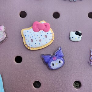 K&ouml;nnte beinhalten: Auswahl an dekorativen Charms mit Cartoon-Charakter-Designs. Enth&auml;lt einen Hello Kitty-Charm mit rosa Schleife, einen lila Kuromi-Charm und andere glitzernde Designs. Die Charms sind in verschiedenen Farben und Stilen.