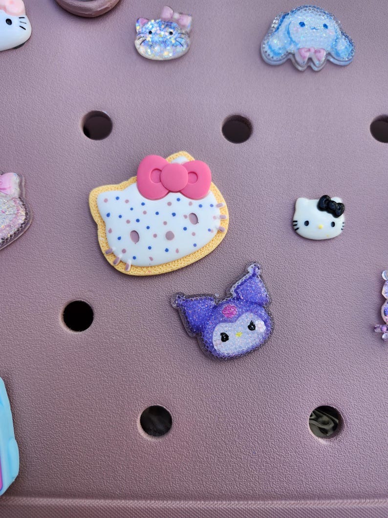 K&ouml;nnte beinhalten: Auswahl an dekorativen Charms mit Cartoon-Charakter-Designs. Enth&auml;lt einen Hello Kitty-Charm mit rosa Schleife, einen lila Kuromi-Charm und andere glitzernde Designs. Die Charms sind in verschiedenen Farben und Stilen.