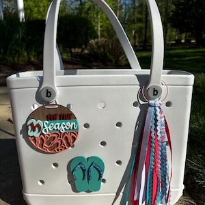 May include: A white tote bag with a pair of flip-flop charms and a tassel. The wooden charm reads "Flip Flop Season" in a layered design with teal and coral accents. The bag has a pair of teal flip-flop stickers and a tassel with red, white, and blue ribbons.