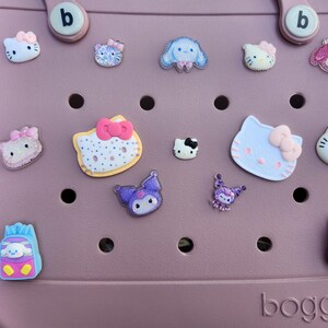 K&ouml;nnte beinhalten: Eine staubrosa Tragetasche mit verschiedenen bunten Charakteranh&auml;ngern. Zu den Anh&auml;ngern geh&ouml;ren Hello Kitty, ein blauer Welpe und andere Zeichentrickfiguren. Die Tasche hat zwei Griffe und das Wort "bogg" ist unten aufgedruckt.