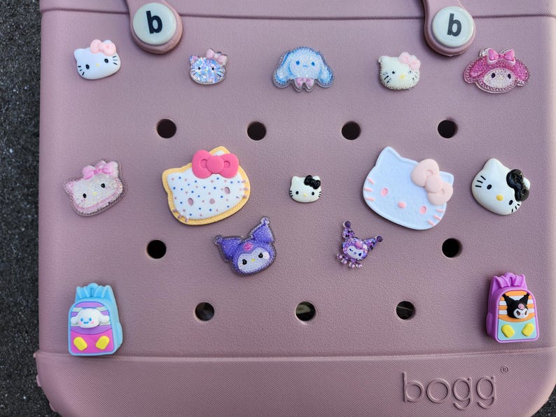 K&ouml;nnte beinhalten: Eine staubrosa Tragetasche mit verschiedenen bunten Charakteranh&auml;ngern. Zu den Anh&auml;ngern geh&ouml;ren Hello Kitty, ein blauer Welpe und andere Zeichentrickfiguren. Die Tasche hat zwei Griffe und das Wort "bogg" ist unten aufgedruckt.