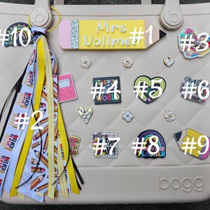 K&ouml;nnte beinhalten: Eine beige Tragetasche mit verschiedenen dekorativen Anh&auml;ngern und einem Quastenanh&auml;nger. Zu den Anh&auml;ngern geh&ouml;ren ein Bleistift mit dem Text "Mrs. Vollme", ein Herz, ein Regenbogen und ein Notizbuch. Der Quastenanh&auml;nger hat gelbe und wei&szlig;e B&auml;nder mit Bleistift- und Textmotiven.