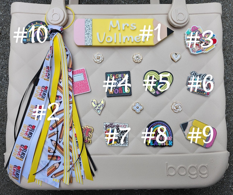 K&ouml;nnte beinhalten: Eine beige Tragetasche mit verschiedenen dekorativen Anh&auml;ngern und einem Quastenanh&auml;nger. Zu den Anh&auml;ngern geh&ouml;ren ein Bleistift mit dem Text "Mrs. Vollme", ein Herz, ein Regenbogen und ein Notizbuch. Der Quastenanh&auml;nger hat gelbe und wei&szlig;e B&auml;nder mit Bleistift- und Textmotiven.