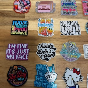 May include: A collection of 14 colorful stickers with various sayings and designs. The stickers feature glitter and a variety of fonts. Some of the sayings include "Have the day you deserve", "I'm fine it's just my face", "On my husband's last nerve", "Normal people scare me", and "I kinda don't care".
