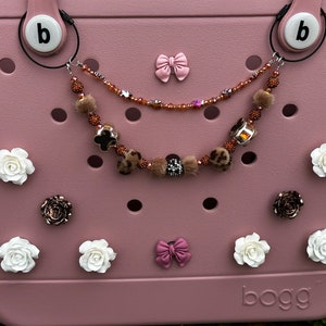 May include: A pink  'BOGG' tote bag with a variety of charms and accessories. The charms include white roses, brown roses, pink bows, and a brown and gold beaded chain with fur pom-poms.