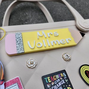 K&ouml;nnte beinhalten: Eine hellbeige Tragetasche mit einem bleistiftf&ouml;rmigen Namensschild mit der Aufschrift "Mrs Vollmer". Das Bild zeigt auch mehrere dekorative Anstecknadeln, darunter ein Notizbuch und eine Anstecknadel mit der Aufschrift "Teachers Can Change The World".