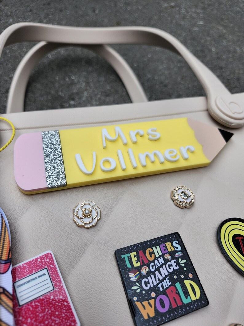 K&ouml;nnte beinhalten: Eine hellbeige Tragetasche mit einem bleistiftf&ouml;rmigen Namensschild mit der Aufschrift "Mrs Vollmer". Das Bild zeigt auch mehrere dekorative Anstecknadeln, darunter ein Notizbuch und eine Anstecknadel mit der Aufschrift "Teachers Can Change The World".