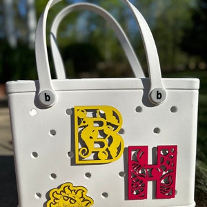 May include: White tote bag with the letters "B" and "H" in yellow and red with cut-out designs. The bag has a "bogg" logo and two white handles.