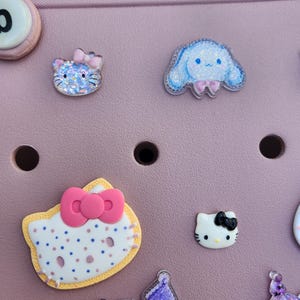 K&ouml;nnte beinhalten: Eine Sammlung farbenfroher, dekorativer Charms mit Zeichentrickfiguren. Die Charms umfassen eine blaue Katze mit rosa Schleife, eine blaue Figur mit rosa Schleife und mehrere Hello Kitty-Designs mit Schleifen. Die Charms befinden sich auf einer rosa Oberfl&auml;che.