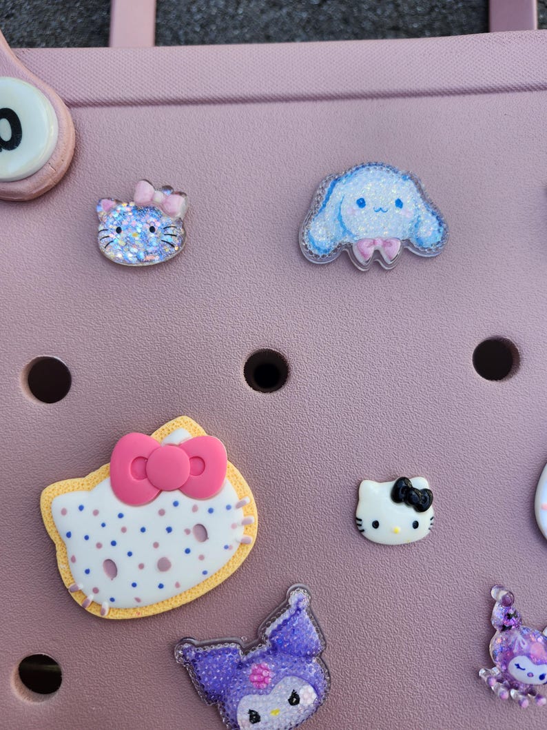 K&ouml;nnte beinhalten: Eine Sammlung farbenfroher, dekorativer Charms mit Zeichentrickfiguren. Die Charms umfassen eine blaue Katze mit rosa Schleife, eine blaue Figur mit rosa Schleife und mehrere Hello Kitty-Designs mit Schleifen. Die Charms befinden sich auf einer rosa Oberfl&auml;che.