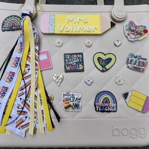 K&ouml;nnte beinhalten: Eine beige Tragetasche, die mit Accessoires zum Thema Lehrer verziert ist. Die Tasche hat ein stiftf&ouml;rmiges Namensschild, dekorative Anstecknadeln und eine Quaste mit B&auml;ndern mit der Aufschrift "Teach, Inspire, Love". Die Tasche hat ein Steppmuster.