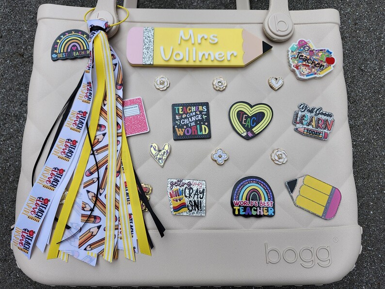 K&ouml;nnte beinhalten: Eine beige Tragetasche, die mit Accessoires zum Thema Lehrer verziert ist. Die Tasche hat ein stiftf&ouml;rmiges Namensschild, dekorative Anstecknadeln und eine Quaste mit B&auml;ndern mit der Aufschrift "Teach, Inspire, Love". Die Tasche hat ein Steppmuster.