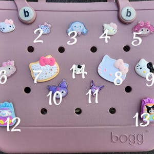 K&ouml;nnte beinhalten: Eine Sammlung dekorativer Charms, darunter Hello Kitty und andere Zeichentrickfiguren, die an einer rosa Tragetasche befestigt sind. Die Charms sind in verschiedenen Farben und Designs gehalten und verleihen der Tasche eine verspielte Note.