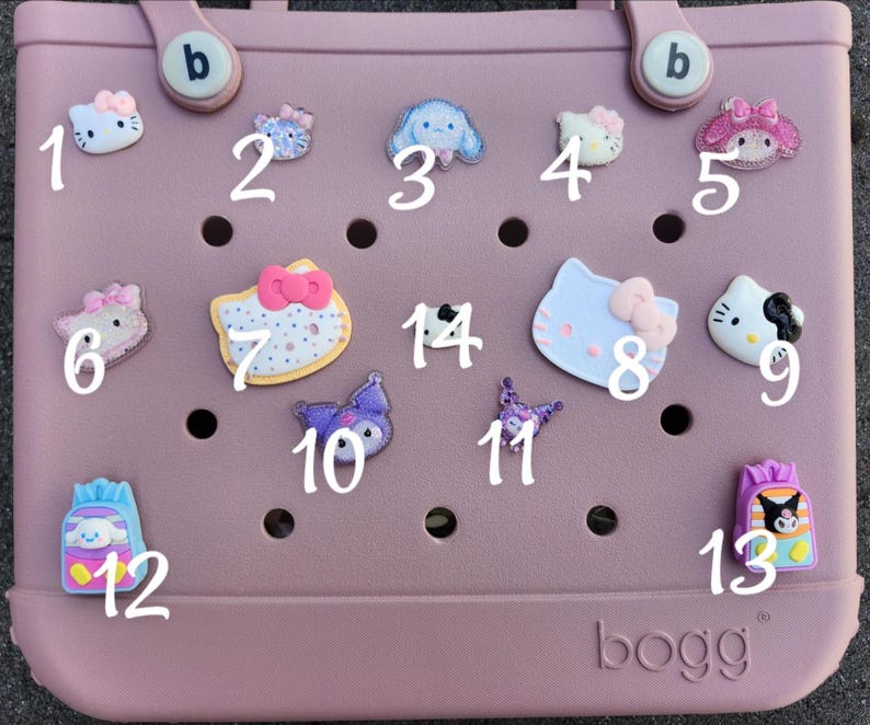 K&ouml;nnte beinhalten: Eine Sammlung dekorativer Charms, darunter Hello Kitty und andere Zeichentrickfiguren, die an einer rosa Tragetasche befestigt sind. Die Charms sind in verschiedenen Farben und Designs gehalten und verleihen der Tasche eine verspielte Note.
