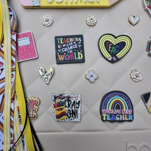 K&ouml;nnte beinhalten: Eine beige Tasche, verziert mit verschiedenen Verzierungen zum Thema Lehrer. Ein gro&szlig;es, gelbes, stiftf&ouml;rmiges Namensschild tr&auml;gt die Aufschrift "Mrs. Vollmer". Weitere Charms sind ein Notizbuch, ein Herz, ein Regenbogen und der Text "Teachers Can Change The World."