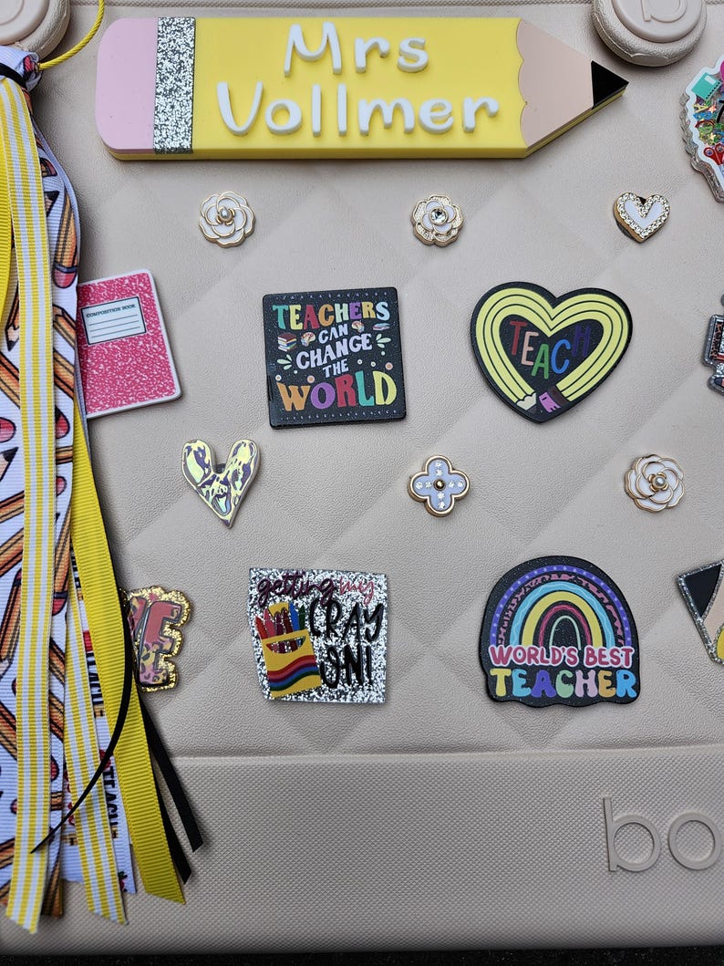 K&ouml;nnte beinhalten: Eine beige Tasche, verziert mit verschiedenen Verzierungen zum Thema Lehrer. Ein gro&szlig;es, gelbes, stiftf&ouml;rmiges Namensschild tr&auml;gt die Aufschrift "Mrs. Vollmer". Weitere Charms sind ein Notizbuch, ein Herz, ein Regenbogen und der Text "Teachers Can Change The World."