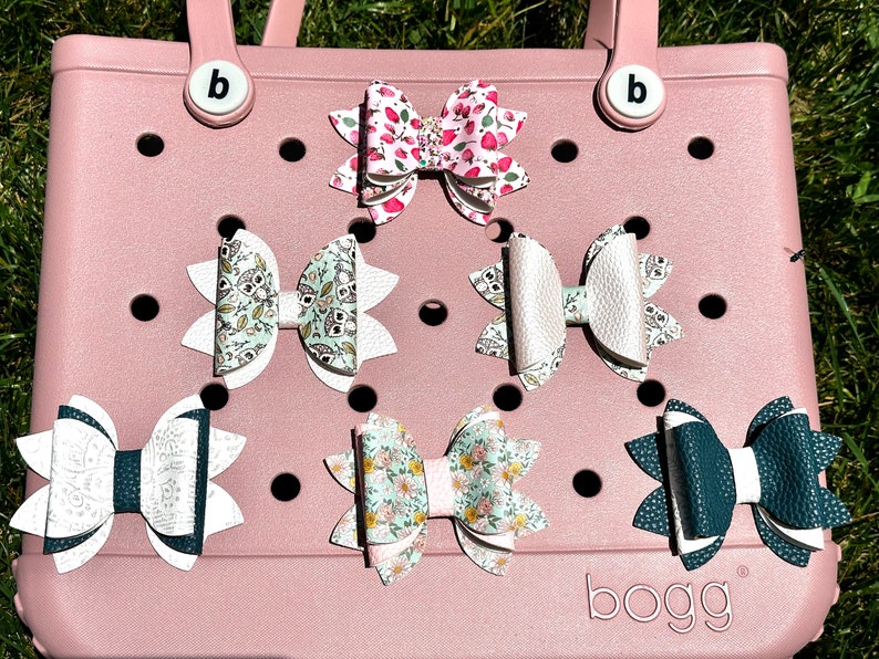 May include: Six different patterned hair bows with white, green, pink, and blue colors. The bows are displayed on a pink, textured surface with the word "bogg" printed on the bottom.