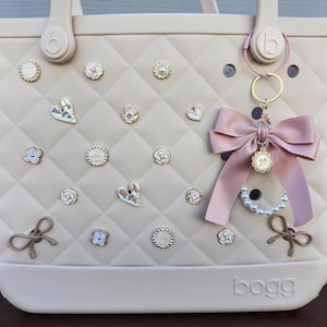 May include: A beige quilted tote bag with decorative embellishments. The bag features a variety of charms, including hearts, flowers, and round accents. A pink bow and pearl strand hang from the handle. The word "bogg" is printed on the bottom.
