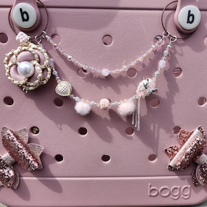 May include: A pink and white  Bogg bag with two pink and gold bows attached to the front. The bag has a charm attached to the top with a pink flower, a pearl, and a silver heart. The charm is attached to a silver chain with pink and white beads.