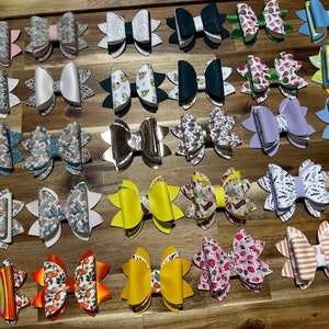 May include: A collection of 25 handmade hair bows in various colors and patterns. The bows are made of fabric and leather and feature a variety of designs, including floral, geometric, and abstract patterns.