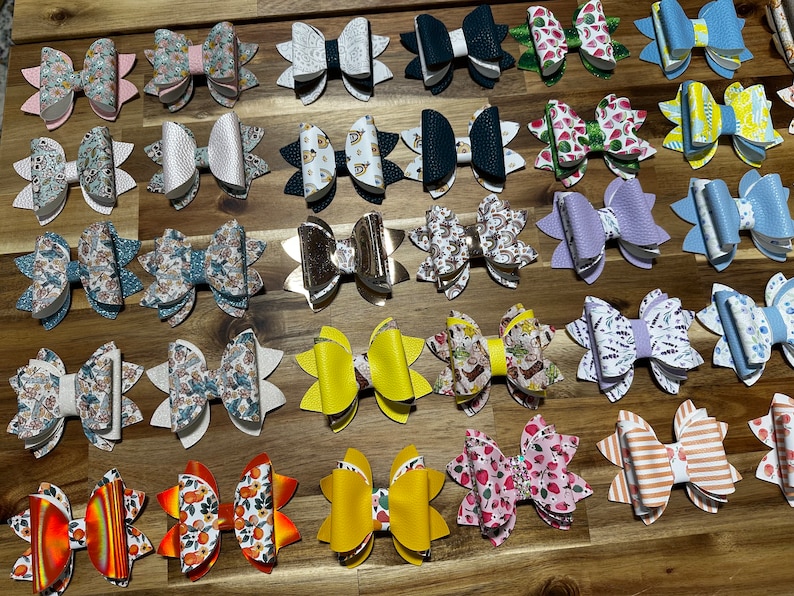 May include: A collection of 25 handmade hair bows in various colors and patterns. The bows are made of fabric and leather and feature a variety of designs, including floral, geometric, and abstract patterns.