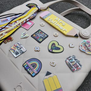 K&ouml;nnte beinhalten: Eine beige Tragetasche mit verschiedenen Lehrer-Themen-Anh&auml;ngern und Accessoires. Zu den Anh&auml;ngern geh&ouml;ren ein Bleistift mit dem Text "Mrs. Vollmer", ein Regenbogen mit "World's Best Teacher" und andere schulbezogene Designs. Die Tasche hat einen Griff und das Wort "boga" an der Seite.
