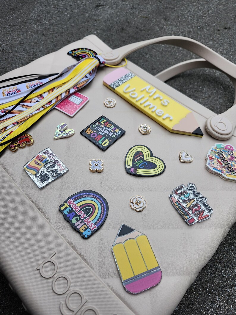 K&ouml;nnte beinhalten: Eine beige Tragetasche mit verschiedenen Lehrer-Themen-Anh&auml;ngern und Accessoires. Zu den Anh&auml;ngern geh&ouml;ren ein Bleistift mit dem Text "Mrs. Vollmer", ein Regenbogen mit "World's Best Teacher" und andere schulbezogene Designs. Die Tasche hat einen Griff und das Wort "boga" an der Seite.