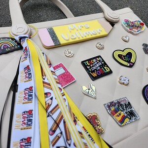 K&ouml;nnte beinhalten: Eine beige Tragetasche mit einem bleistiftf&ouml;rmigen Namensschild mit der Aufschrift "Mrs. Vollmer". Die Tasche ist mit verschiedenen Lehrer-Themen-Anh&auml;ngern verziert, darunter Herzen, Blumen und ein Anh&auml;nger mit der Aufschrift "Teachers Can Change the World". Ein Quaste mit B&auml;ndern mit der Aufschrift "Teach, Love, Inspire" ist befestigt.