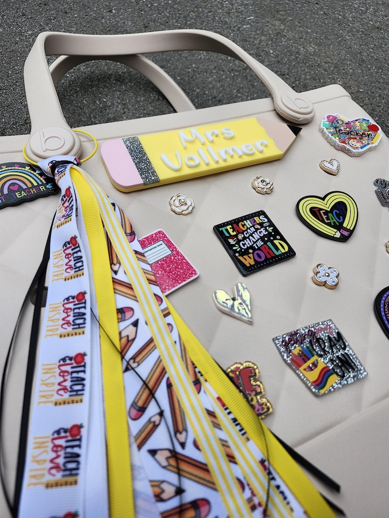 K&ouml;nnte beinhalten: Eine beige Tragetasche mit einem bleistiftf&ouml;rmigen Namensschild mit der Aufschrift "Mrs. Vollmer". Die Tasche ist mit verschiedenen Lehrer-Themen-Anh&auml;ngern verziert, darunter Herzen, Blumen und ein Anh&auml;nger mit der Aufschrift "Teachers Can Change the World". Ein Quaste mit B&auml;ndern mit der Aufschrift "Teach, Love, Inspire" ist befestigt.